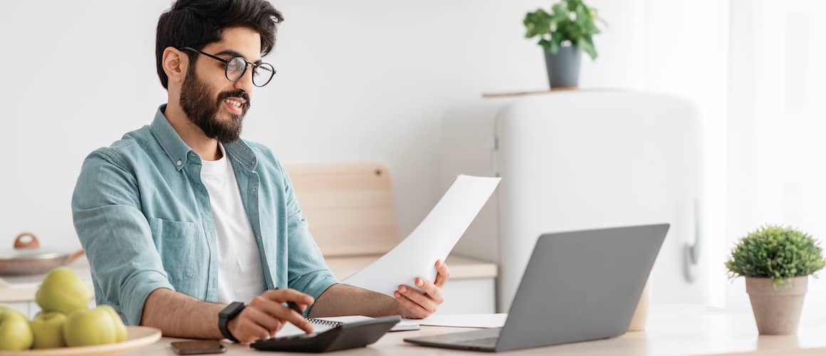 Young adult male with glasses and beard reviewing document and using calculator while sitting in front of open laptop. 