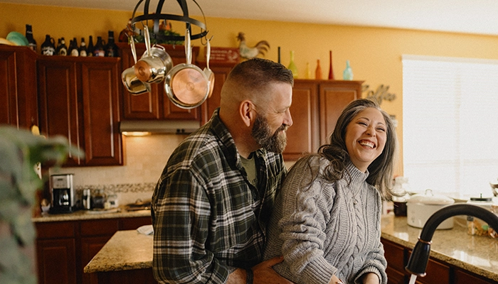 Man holding woman while she does dishes and laughs.