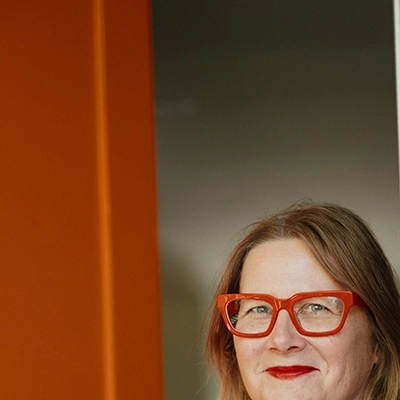 Woman with red glasses and red lips holding open orange door and smiling.