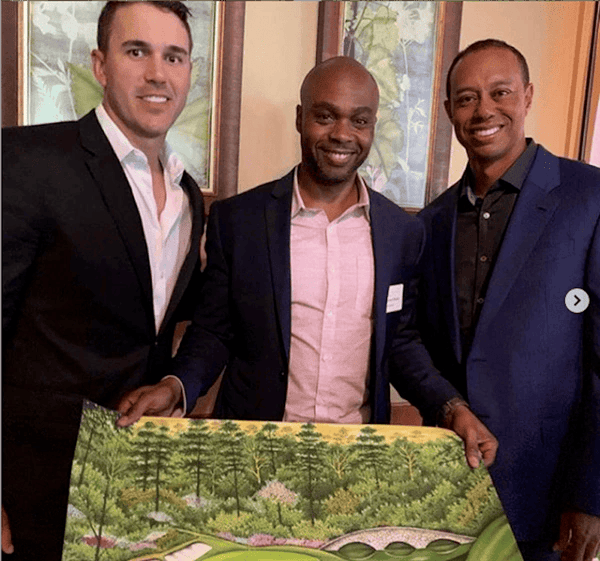 Three men smiling, one is holding a print of a golf course he painted. 