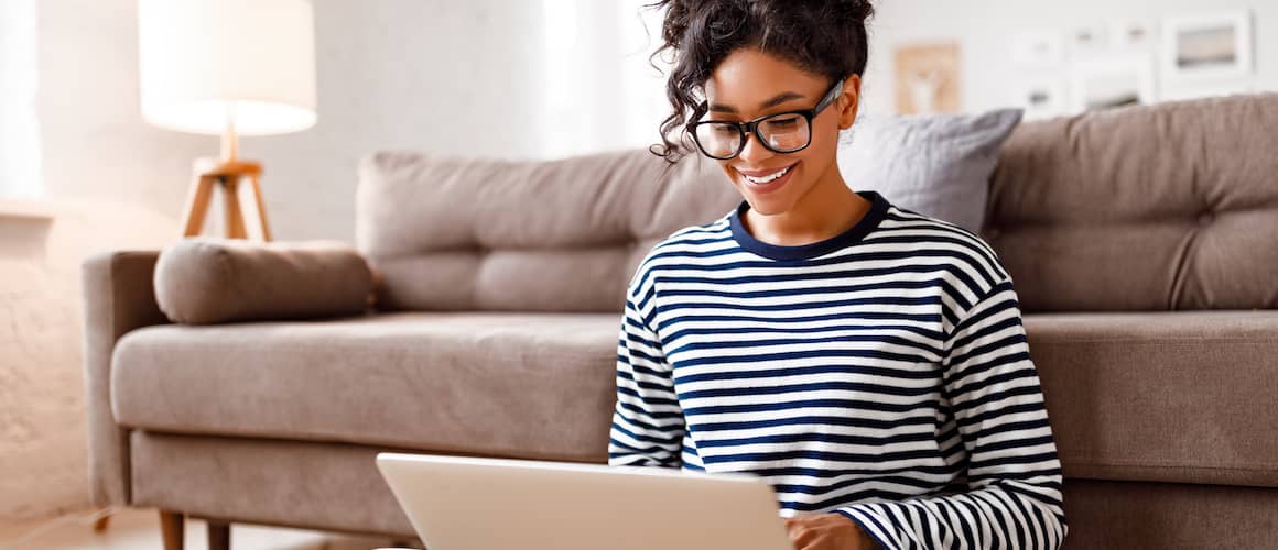 Image of woman sitting next to couch, smiling down at laptop