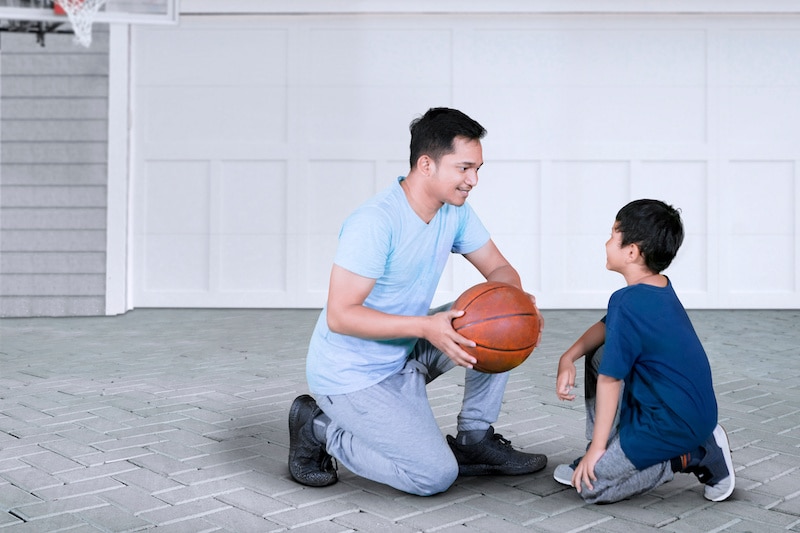 RHB Assets From IGX: Father teaching son to play basketball in a sunlit driveway.