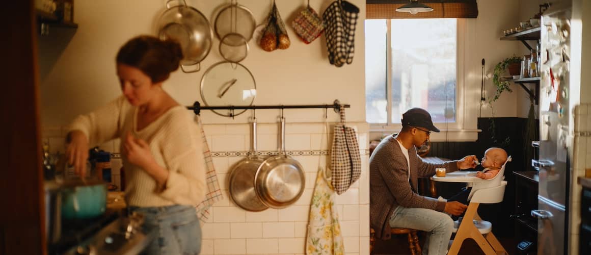 Mother in kitchen cooking while father feeds child in high chair in dining room.