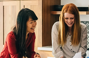 A young Asian woman and her partner, a young white woman with auburn hair, go over home buying plans at their kitchen counter together.
