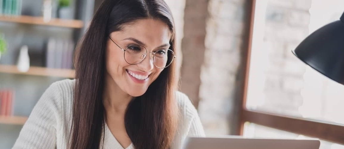 A lady with glasses looking extremely happy while using her laptop.
