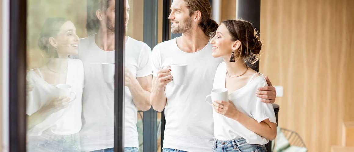 Young couple smiling, having coffee while standing looking out large picture window of new home.