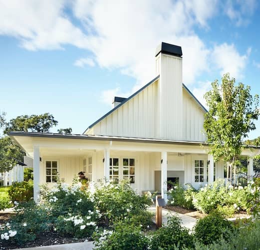 White house with large wrap around covered porch and lush landscaping.