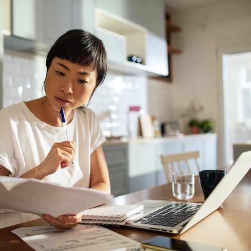Woman in kitchen reviewing paperwork with open laptop nearby.