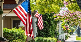 American flags hanging in front of homes with green trees and pink flowers.