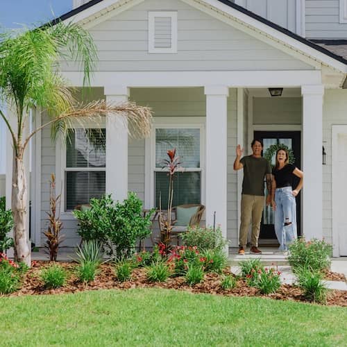 A young couple waving from the front porch of their home.