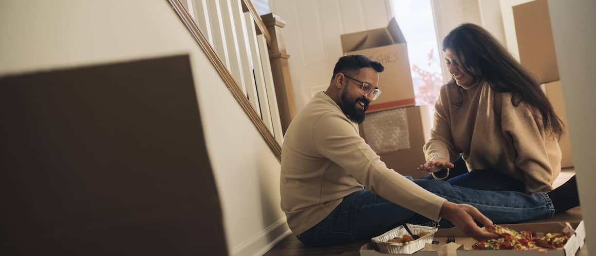 Couple smiling and eating pizza on floor of new home surrounded by unopened boxes.