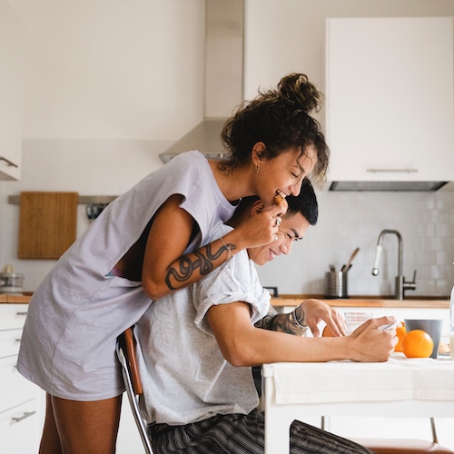 Young couple in kitchen looking at tablet together. There is an orange on the kitchen table nearby.