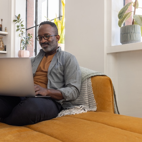 Man on laptop, smiling, sitting on chaise lounge couch.
