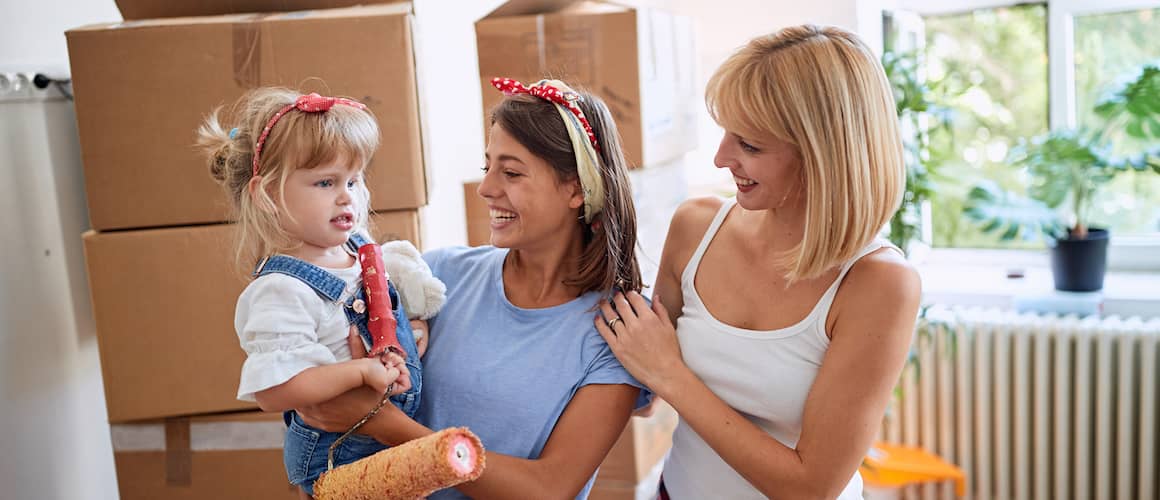 Young family with two moms and a young girl stand smiling in front of large stacks of packed boxes.