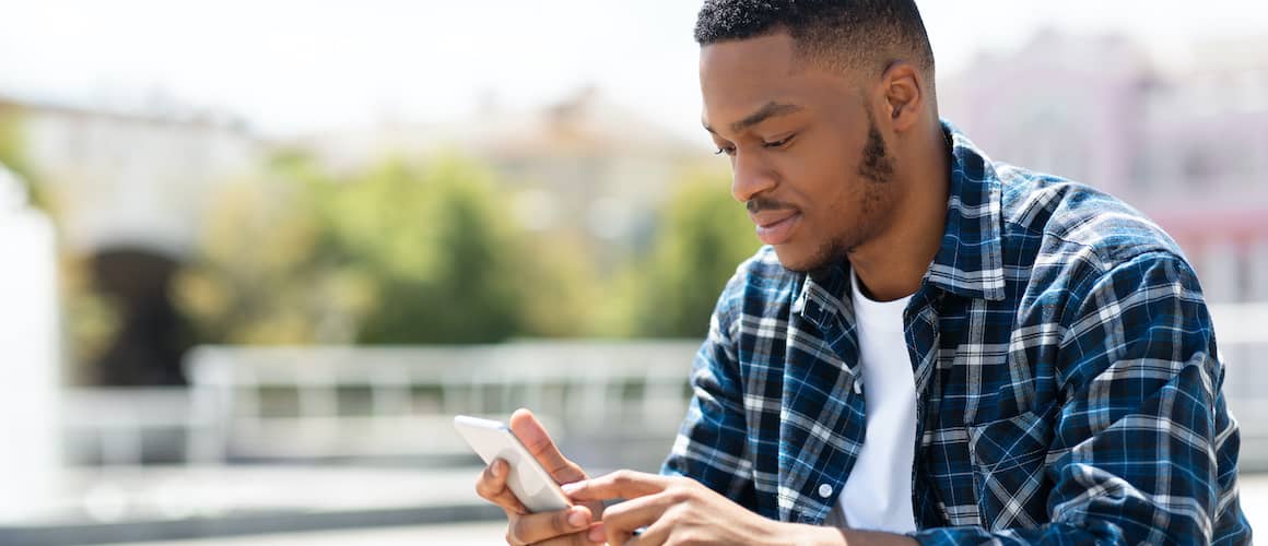 A black man in a blue plaid shirt looking at his phone on a sunny day.