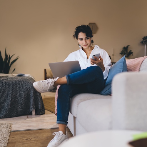 Young woman smiling and sitting on a couch while listening to music on her phone and using her laptop.