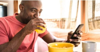 A man drinks a cup of orange juice while reading an article on his phone. 